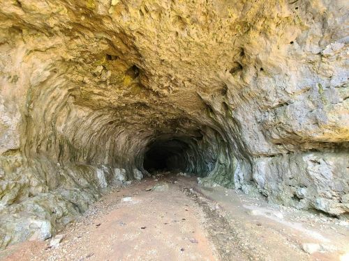 A rocky cave entrance with textured walls and a sandy floor, leading into darkness.