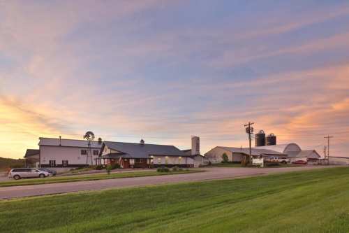 A scenic farm at sunset, featuring a white farmhouse, silos, and green fields under a colorful sky.