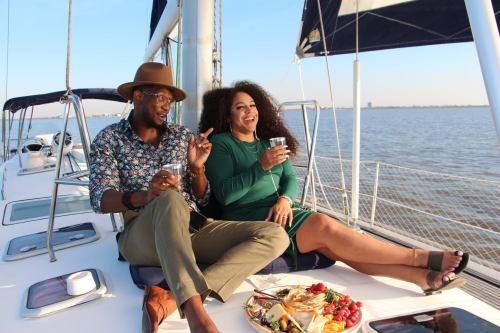 A couple enjoys drinks on a sailboat, smiling and relaxing by the water with a platter of food nearby.