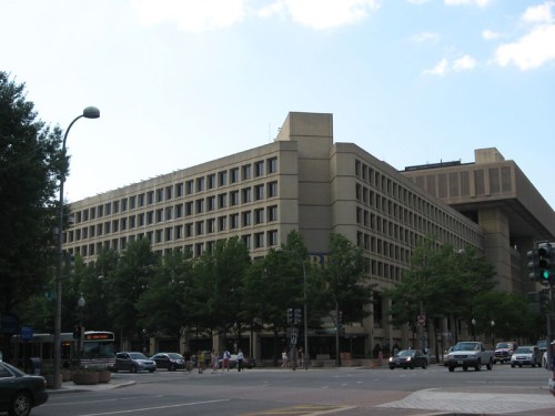 A large, modern building with multiple windows, surrounded by trees and a busy street with cars and pedestrians.