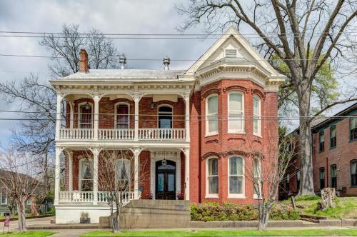 Victorian-style brick house with a wraparound porch, large windows, and a tree in the yard under a cloudy sky.