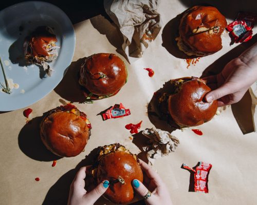 A top-down view of several burgers on brown paper, with hands reaching for them and scattered ketchup packets.