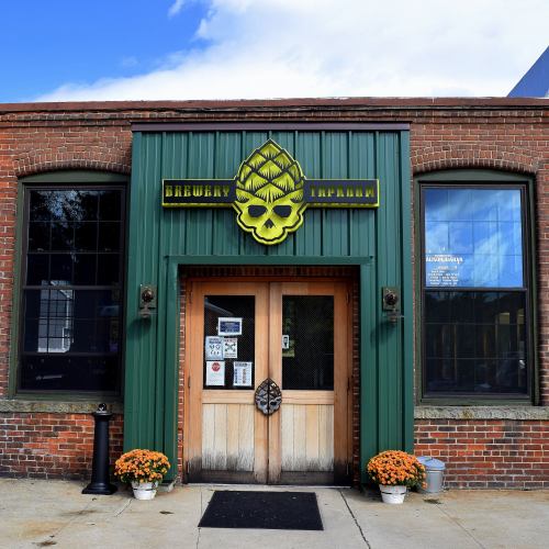 Exterior of a brewery with a green and yellow sign featuring a skull and hop design, surrounded by brick walls and flowers.