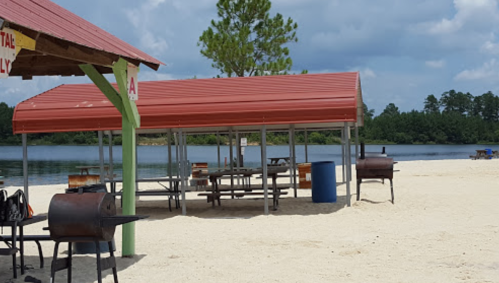 Beach scene with red-roofed pavilions, picnic tables, and grills near a calm lake under a partly cloudy sky.