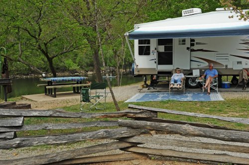 A camper by a river with two people relaxing in chairs, surrounded by trees and a rustic wooden fence.