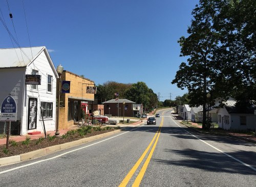 A quiet street scene with shops and a car driving, under a clear blue sky. Green trees line the road.