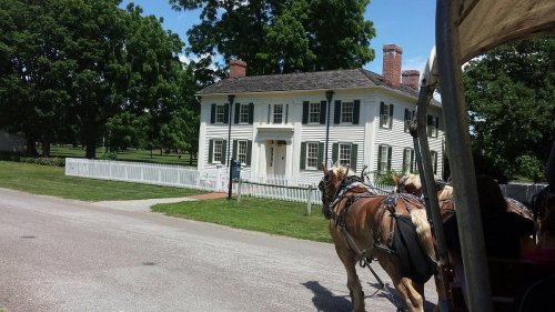A historic white house with green shutters, seen from a horse-drawn carriage on a sunny day. Lush trees surround the area.