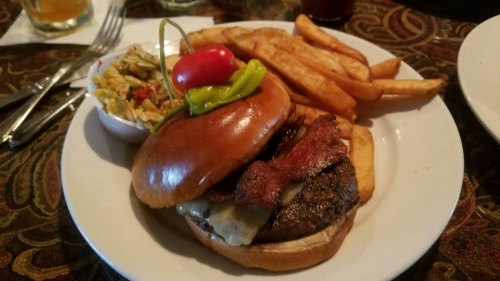 A juicy burger topped with bacon and cheese, served with fries and a side of coleslaw, garnished with a cherry tomato.