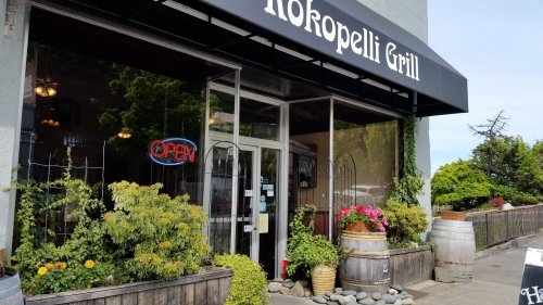 Exterior of Hokopelli Grill with an "Open" sign, surrounded by greenery and colorful flowers.