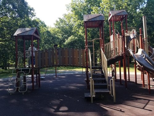 A playground with slides and climbing structures surrounded by trees on a sunny day. Two children are playing.