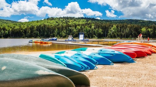 Colorful kayaks lined up on a sandy beach by a calm lake, surrounded by lush green trees and a blue sky with clouds.