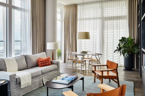 Bright, modern living room with large windows, a cozy sofa, wooden chairs, and a dining table, featuring light curtains.