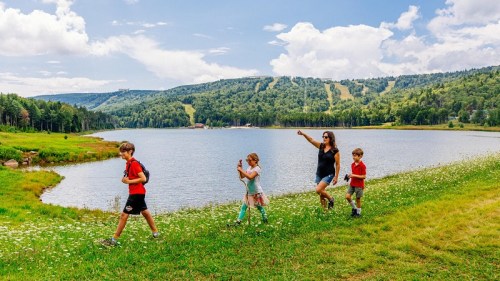 A woman and three children walk along a grassy path by a lake, surrounded by trees and hills under a blue sky.