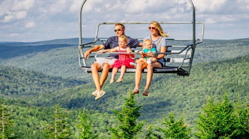 A family of four enjoys a scenic chairlift ride over lush green mountains on a sunny day.