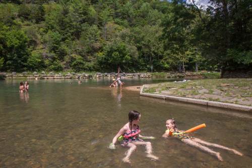 Two children play in a shallow river, while others relax and swim nearby in a lush, green setting.