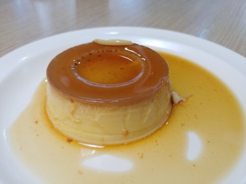 A smooth, round flan topped with caramel sauce, sitting on a white plate with a light wooden background.