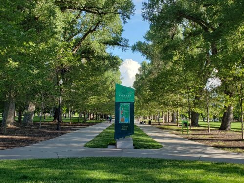 A pathway lined with trees leads to a sign in Liberty Park, under a blue sky with fluffy clouds.