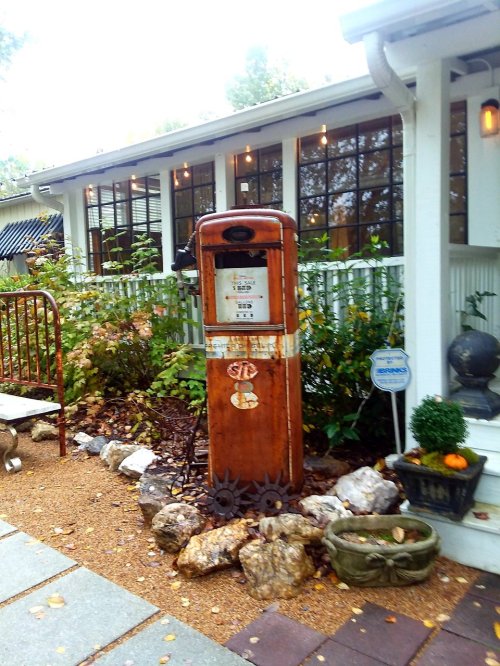 Vintage gas pump surrounded by rocks and plants, near a white house with large windows and outdoor seating.