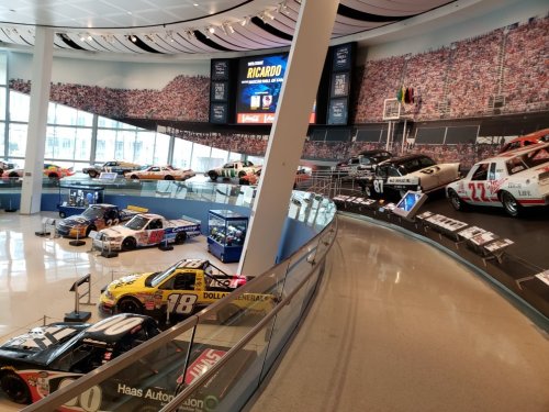 A museum display featuring various race cars arranged along a curved wall with a large crowd mural in the background.