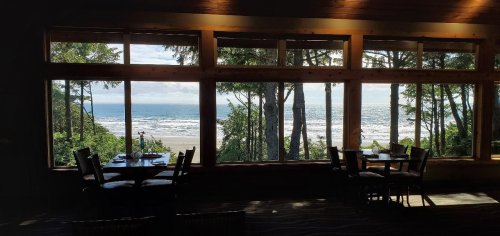 A serene view of the ocean through large windows, framed by trees and a cozy dining area with tables.