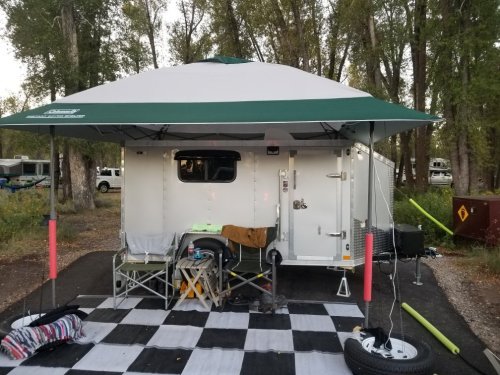 A camping trailer with a green canopy set up in a wooded area, featuring outdoor furniture and a checkered mat.