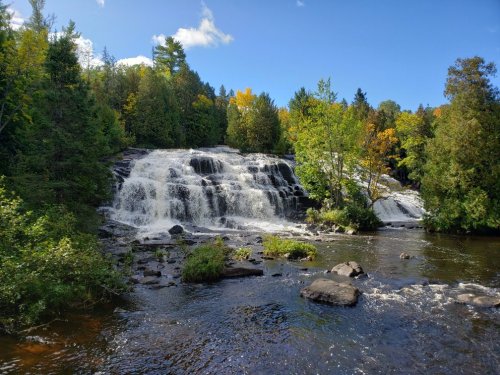 A serene waterfall cascades over rocks, surrounded by lush green trees and autumn foliage under a clear blue sky.