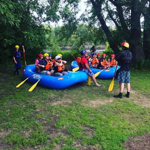 A group of people in life jackets and helmets gather around blue rafts, preparing for a rafting adventure by a river.