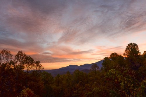 A serene sunset over mountains, with colorful clouds and trees in the foreground, creating a peaceful landscape.