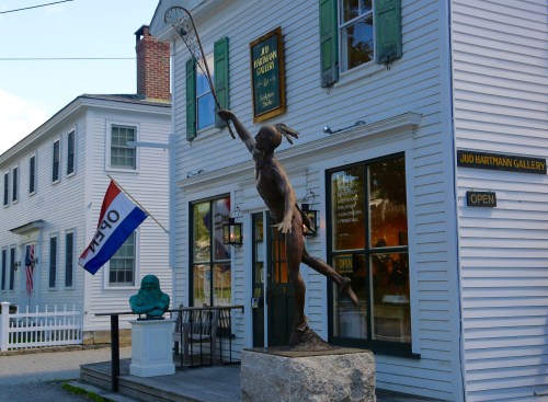 A bronze statue of a tennis player stands outside the Jud Hartmann Gallery, with a flag and buildings in the background.