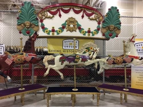 A vintage carousel with three ornate horses, decorated with flowers and flags, displayed behind a fence.