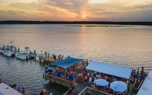 A lively waterfront scene at sunset, with people dining on a dock and boats moored nearby.