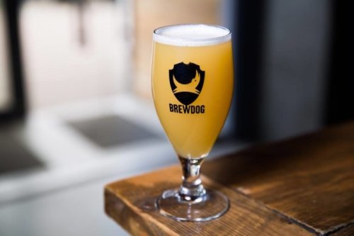 A glass of BrewDog beer sits on a wooden table, showcasing a light golden color and a frothy top.