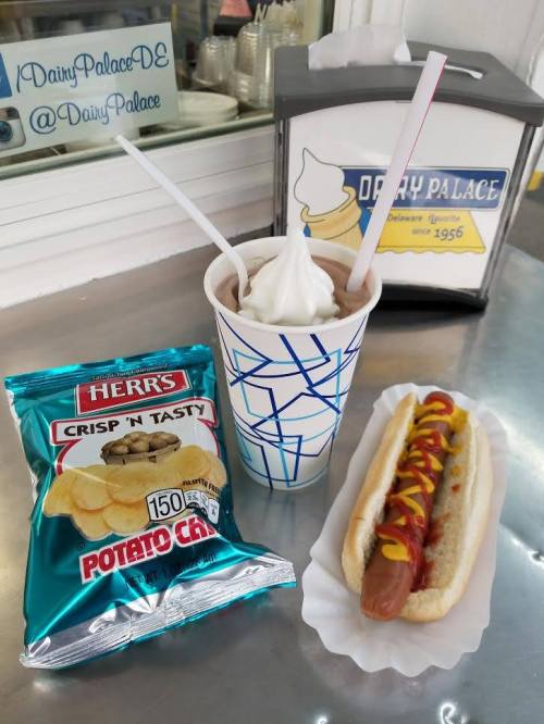 A hot dog with mustard, a cup of soft serve ice cream, and a bag of Herr's potato chips on a table.