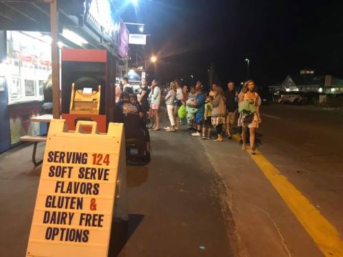 A crowd gathers outside a soft serve ice cream shop at night, with a sign highlighting 124 flavors and gluten/dairy-free options.