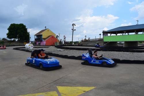 Two go-karts race on a track, with one driver wearing a hat and a passenger in the first kart. Colorful buildings in the background.