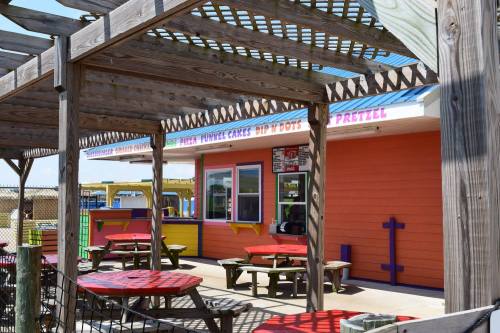Colorful food stand with outdoor seating under a wooden pergola, featuring signs for snacks like funnel cakes and pretzels.