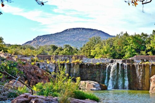 A serene waterfall cascades over rocks, surrounded by lush greenery and a distant mountain under a clear blue sky.