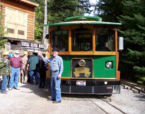 A green trolley bus parked near a group of people in hard hats, surrounded by trees and a wooden building.