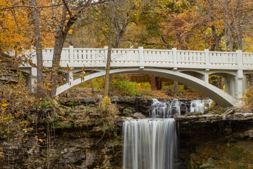 A serene waterfall flows beneath a white arch bridge, surrounded by autumn foliage in a tranquil forest setting.