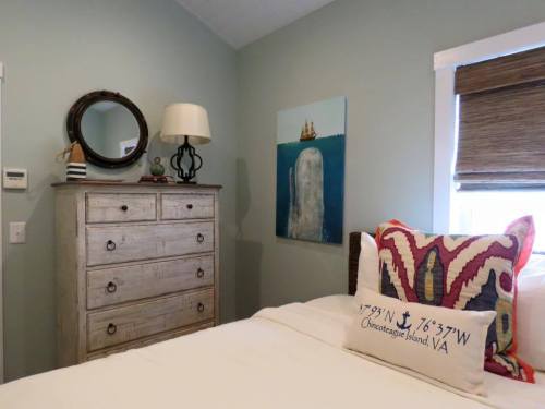 Cozy bedroom with a wooden dresser, lamp, artwork of a ship, and decorative pillows on a bed.