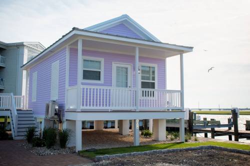 A pastel purple beach house on stilts, featuring a porch and surrounded by gravel and grass, near a waterfront.