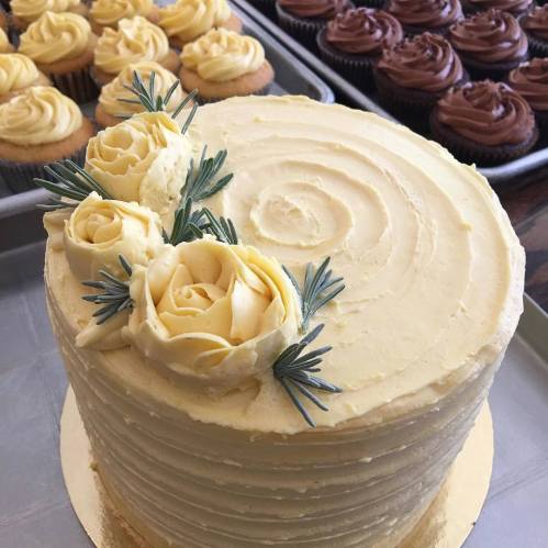 A beautifully decorated yellow cake with buttercream roses and rosemary, surrounded by chocolate and vanilla cupcakes.