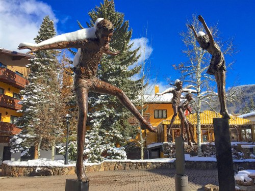 Three bronze sculptures of ice skaters in a snowy outdoor setting, with trees and buildings in the background.