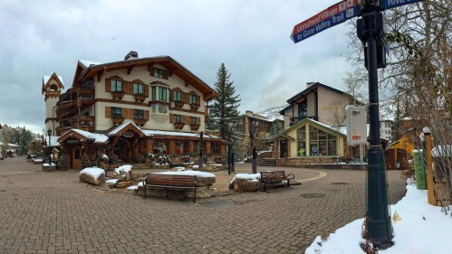 A snowy village square with charming buildings, benches, and trees, under a cloudy sky. Signs point to nearby trails.