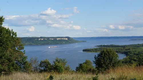 A scenic view of a river with lush green banks, cliffs in the distance, and a boat navigating the water under a blue sky.
