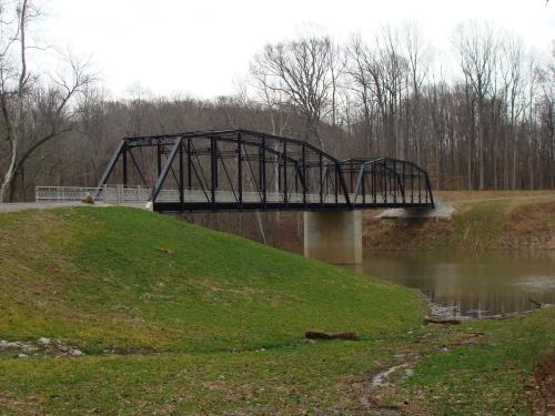 A black metal bridge spans a calm river, surrounded by green grass and bare trees in a serene landscape.