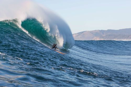 A surfer rides a large wave, showcasing skill and balance against a backdrop of rolling ocean and distant hills.