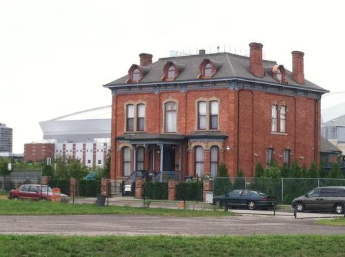 A large, historic red brick house with a gray roof, surrounded by greenery, near modern buildings in the background.