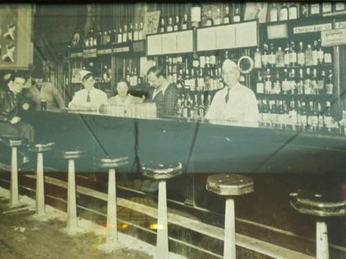A vintage black-and-white photo of a bar with patrons and a bartender, showcasing a long counter and shelves of bottles.