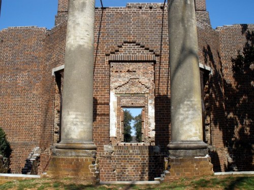 Ruins of a brick building with two large columns and a broken window, surrounded by greenery under a clear blue sky.